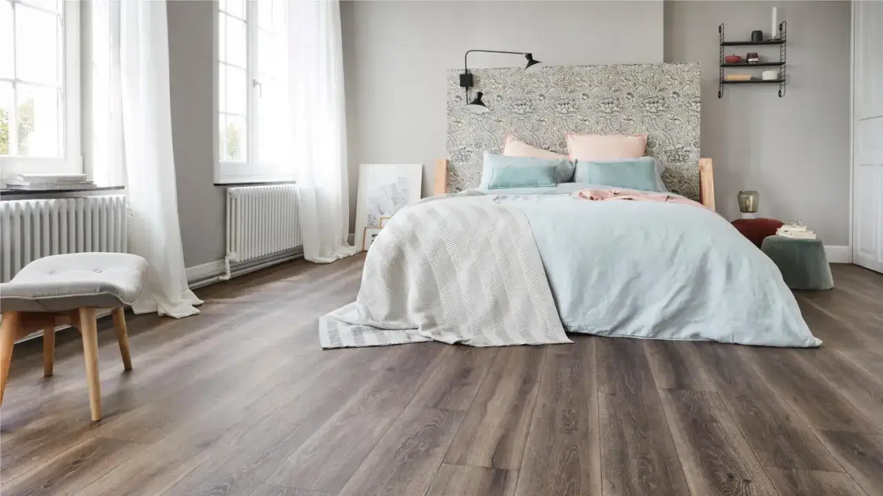 Un lit douillet avec un couvre-lit ray&eacute; et des coussins bleus et roses, sur un **rev&ecirc;tement sol chambre** imitation bois sombre.