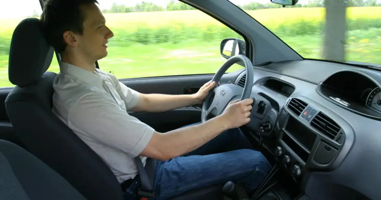 Mężczyzna w jasnej koszuli i jeansach prowadzi auto. Widać jego ręce na kierownicy i fragment deski rozdzielczej. To jak ruszyć autem w praktyce.