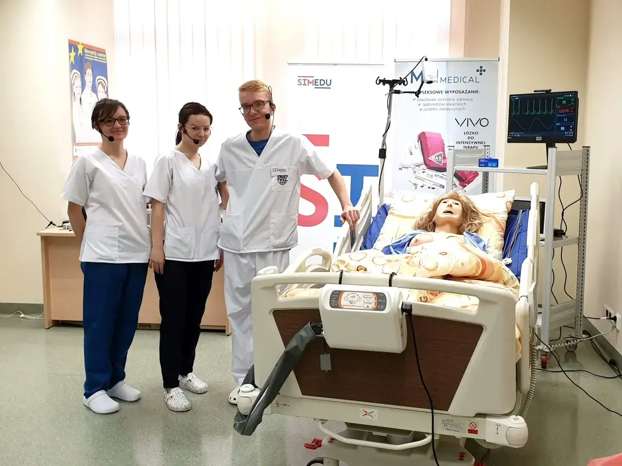 Trzech studentów medycyny w białych fartuchach podczas kursów dla pielęgniarek, obok łóżka symulacyjnego z manekinem.