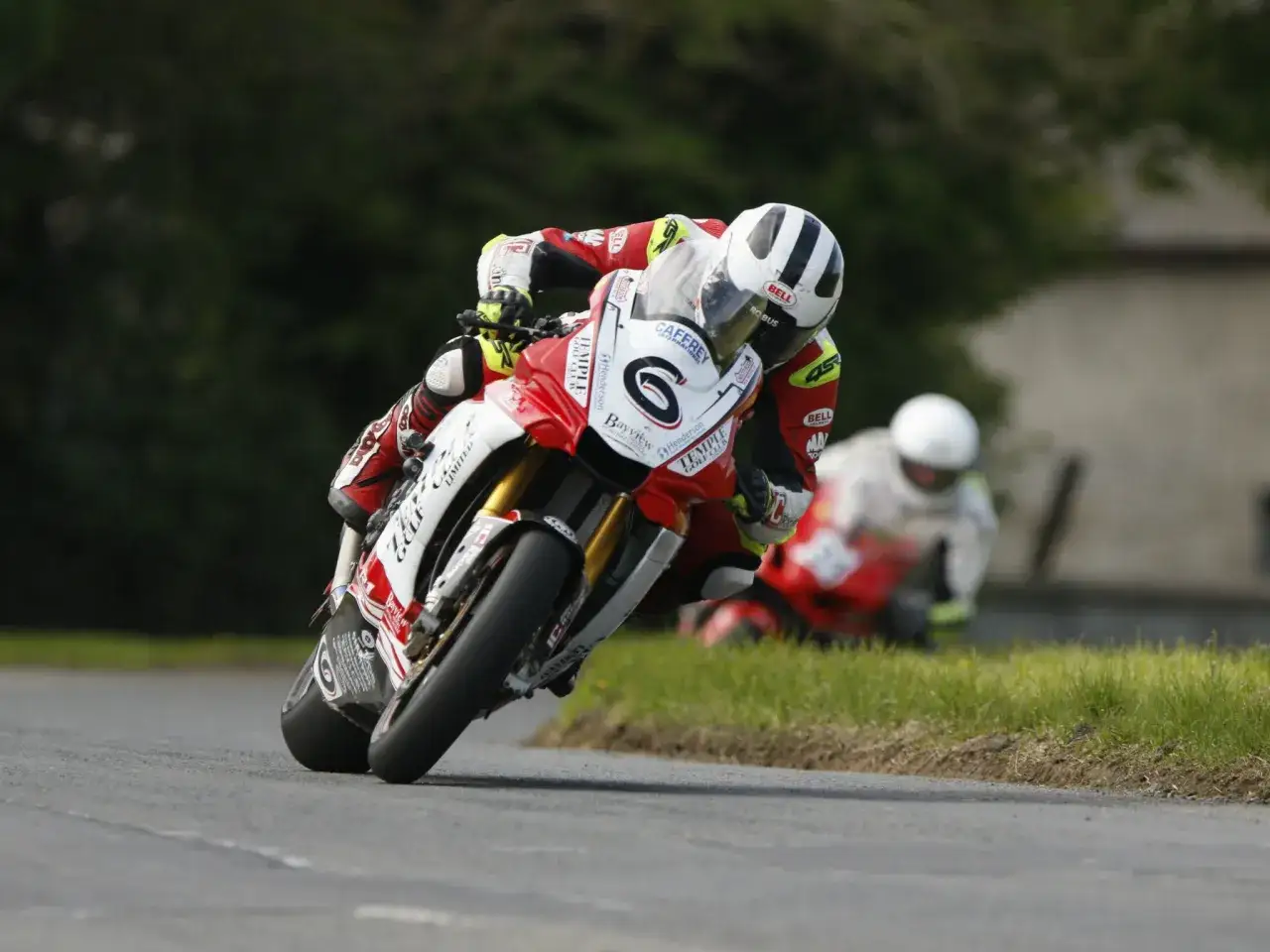 William Dunlop inclinado en su moto de carreras roja y blanca, compitiendo en una carretera con otro corredor detrás.