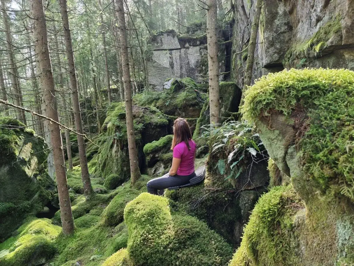 En kvinna vilar på en mossig sten under sin vandring på Bohusleden. Skogen är grön och klipporna täckta av mjuk mossa.