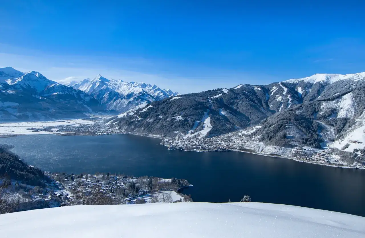Zell am See zimową porą: ośnieżone góry, ciemny, spokojny akwen i urokliwe miasteczko w dole.