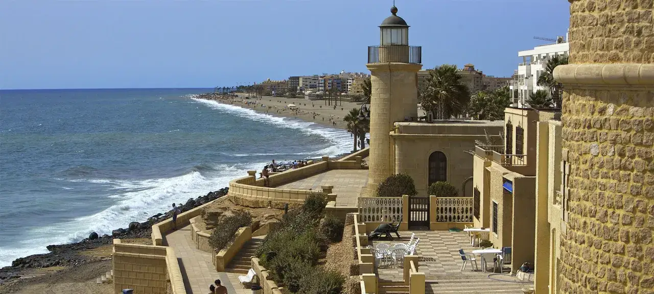 Vista de la costa con un faro, playa con gente y edificios. Un día perfecto para ver roquetas de mar.