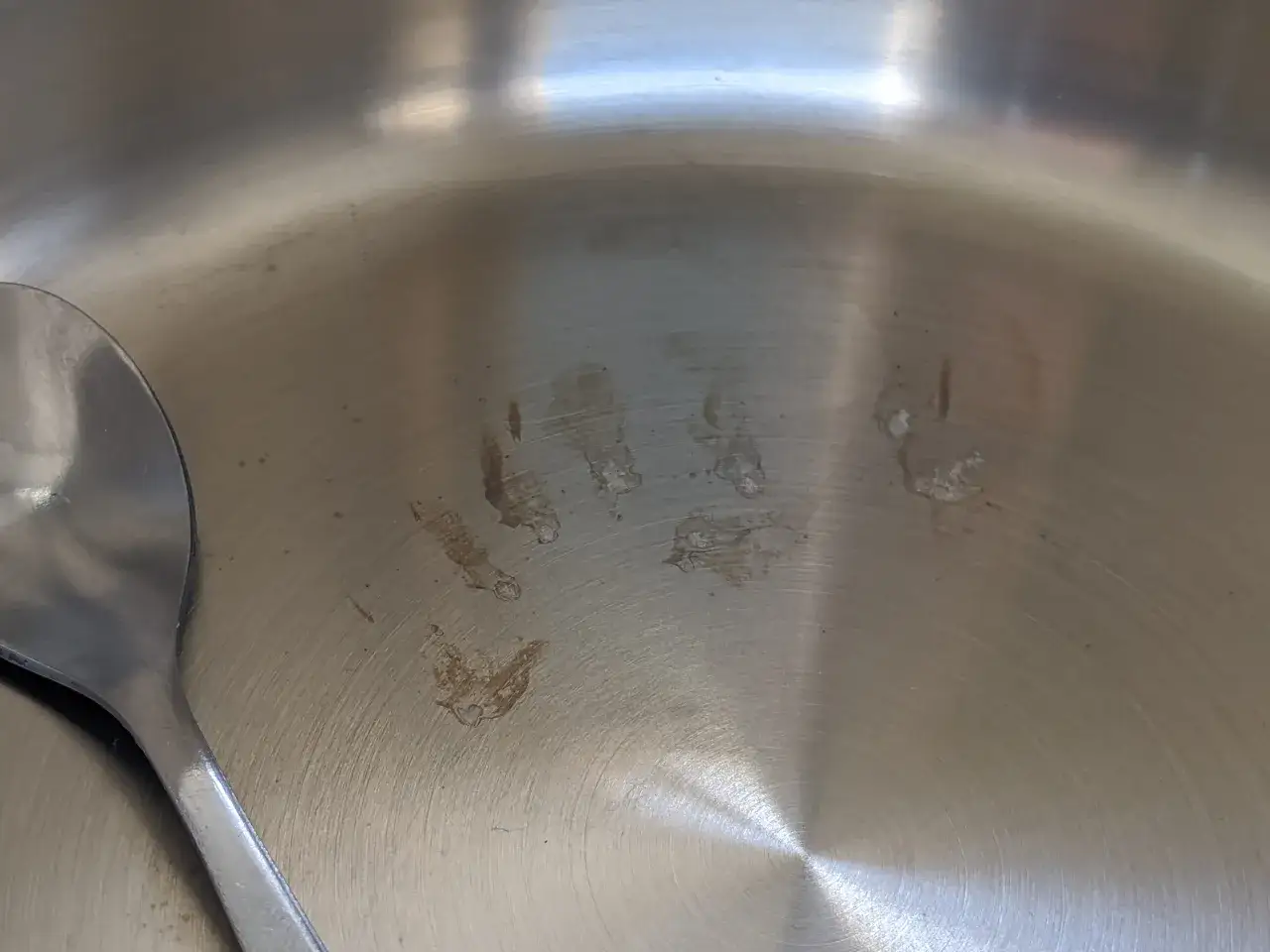 Une cuillère repose dans une casserole en inox, marquée de quelques taches bleues et de résidus de cuisson.