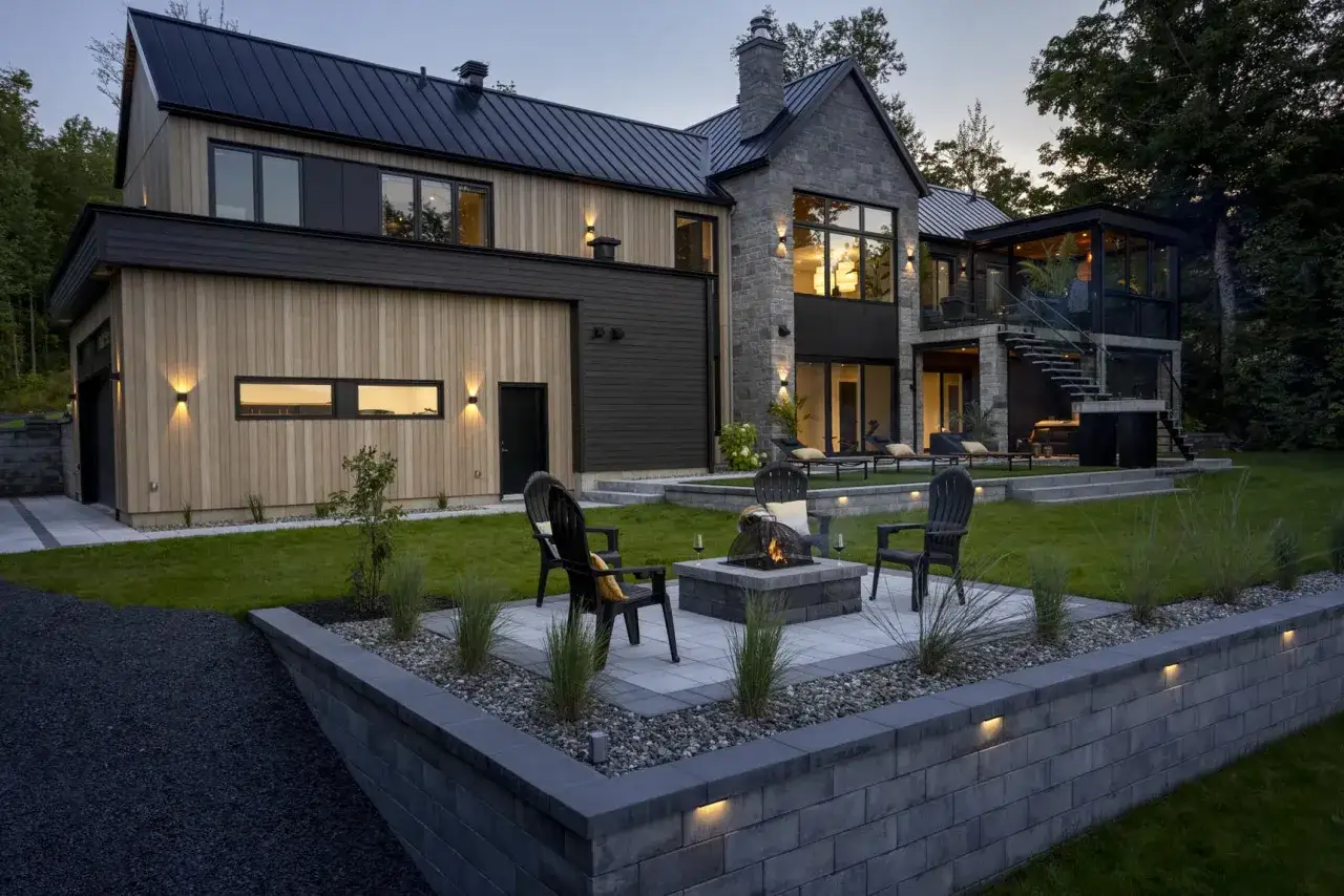 Modern house with stone and wood facade, featuring a fire pit area with chairs on tiered **plots suelo** and a lush green lawn.