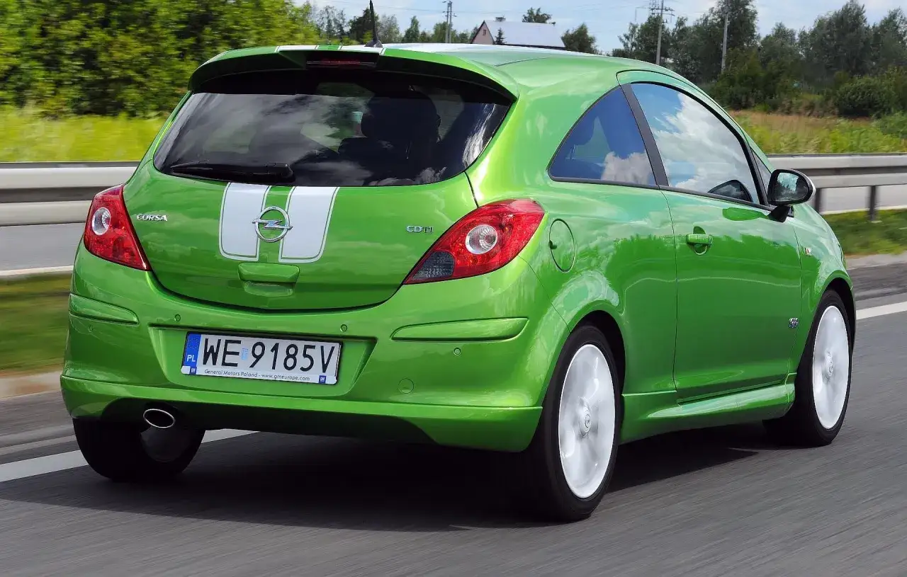 Zielony Opel Corsa D 1.2 benzyna z białymi pasami na klapie bagażnika pędzi po drodze.