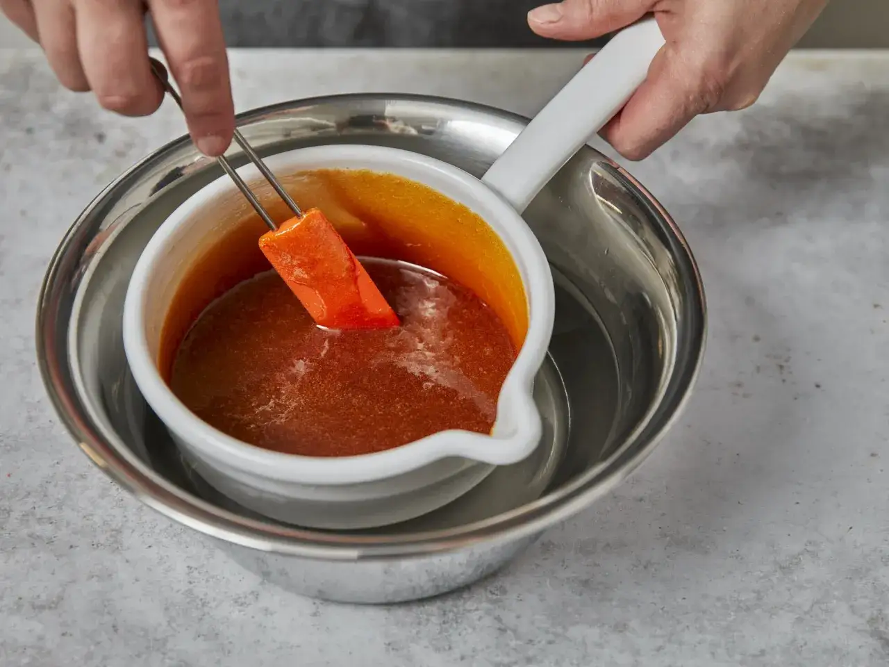 Ein Stück orangefarbener Frucht wird mit einer Zange in eine Schüssel mit karamellisierendem Zucker getaucht.