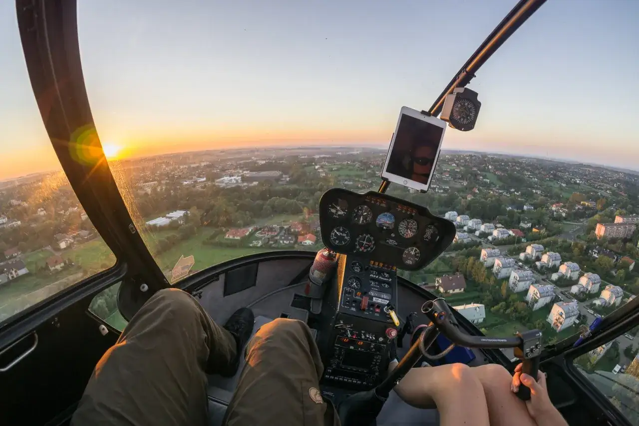 Widok z kokpitu helikoptera o zachodzie słońca. Widoczne nogi pilota, deska rozdzielcza i krajobraz. Marzysz, jak zostać pilotem helikoptera?
