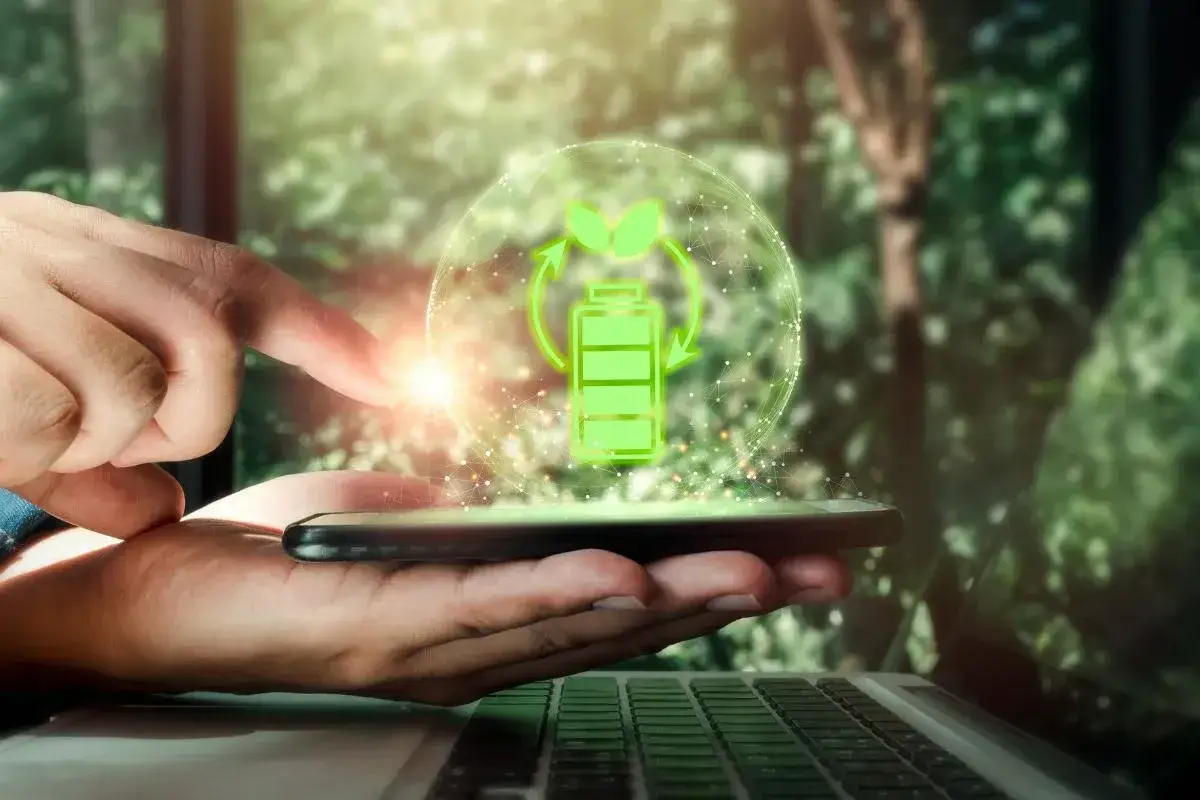 Dłoń trzyma smartfon, nad kt&oacute;rym unosi się zielona ikona baterii z liśćmi i strzałkami, symbolizująca ekologiczne zużycie energii. Zastanawiasz się, ile prądu zużywa komputer?