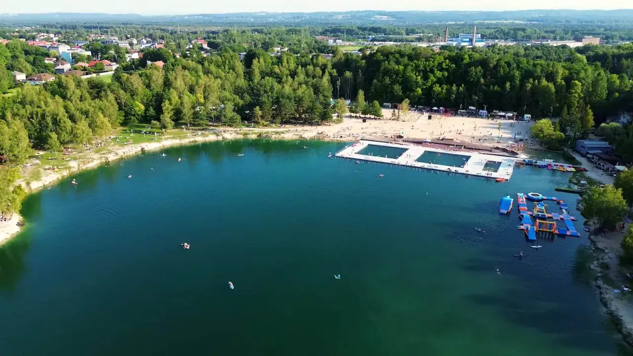 Letni dzień nad wodą w Trzebini. Ludzie odpoczywają na plaży i pływają w jeziorze, które przypomina Balaton. Widać też baseny i dmuchane atrakcje wodne.