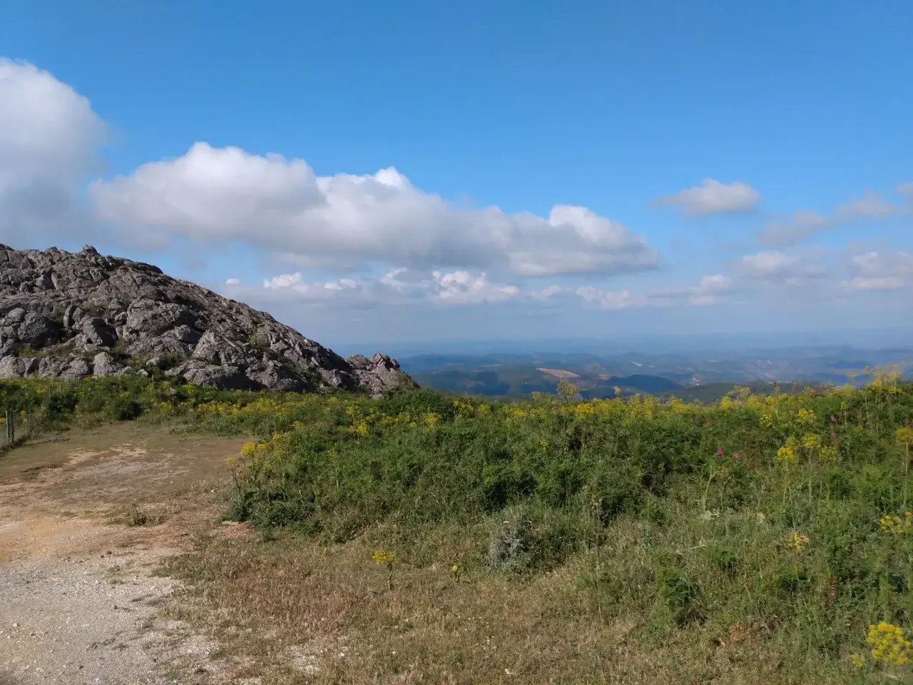 Felsen und bl&uuml;hende Wiesen auf dem Weg nach Monchique, Portugal, unter einem blauen Himmel mit Wolken.