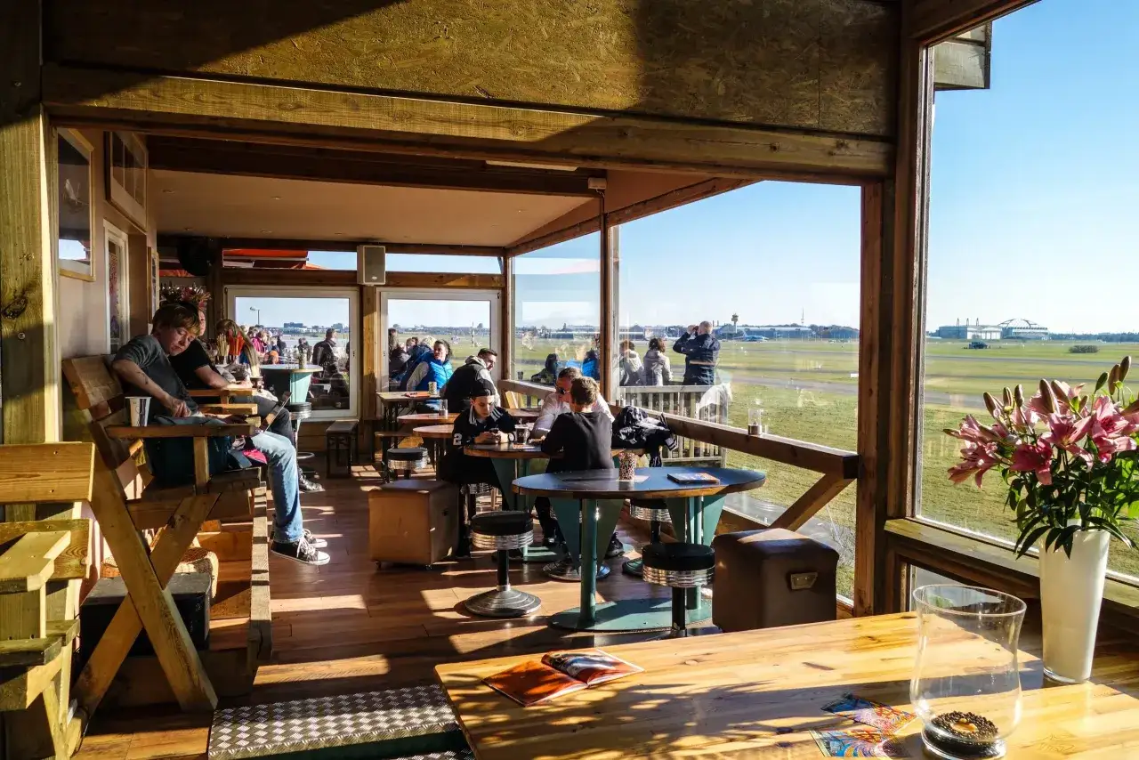 Gemütliches **Cafe to fly** mit Blick auf das Flugfeld. Gäste entspannen sich, während Flugzeuge starten und landen.