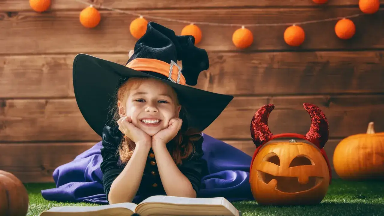 Ni&ntilde;a bruja sonriente lee un libro, rodeada de calabazas y luces de Halloween. Fomenta la comprensi&oacute;n lectora en esta festividad.