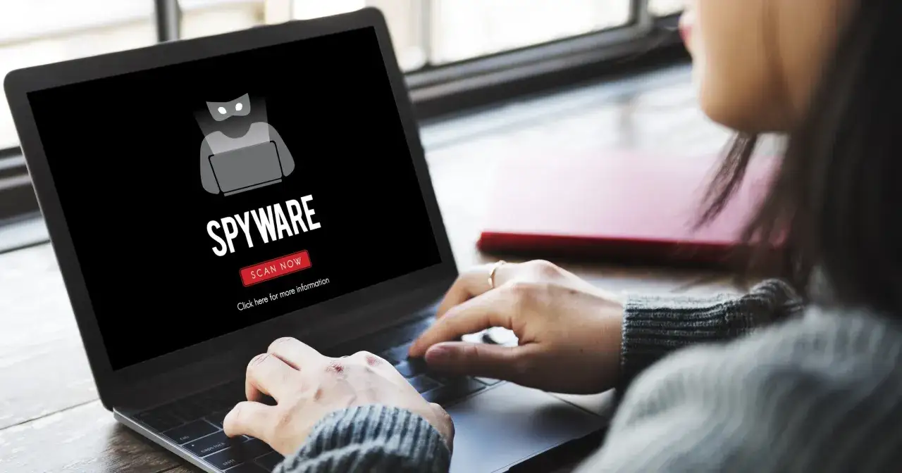 Osoba pracuje na laptopie, na ekranie którego widnieje ostrzeżenie o **spyware** i przycisk 