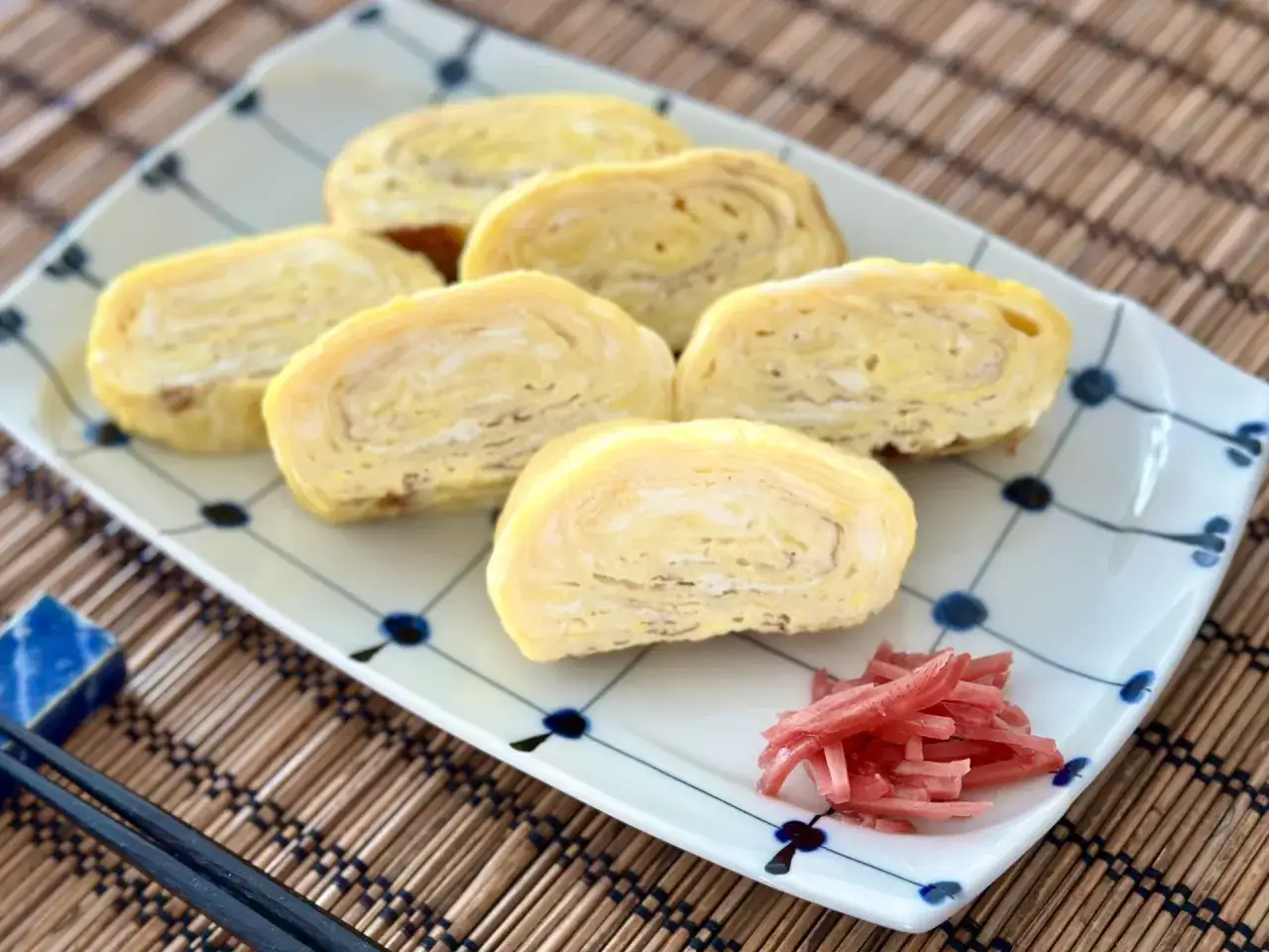 Sześć kawałków omletu tamago ułożonych na talerzu, obok różowy imbir i pałeczki.