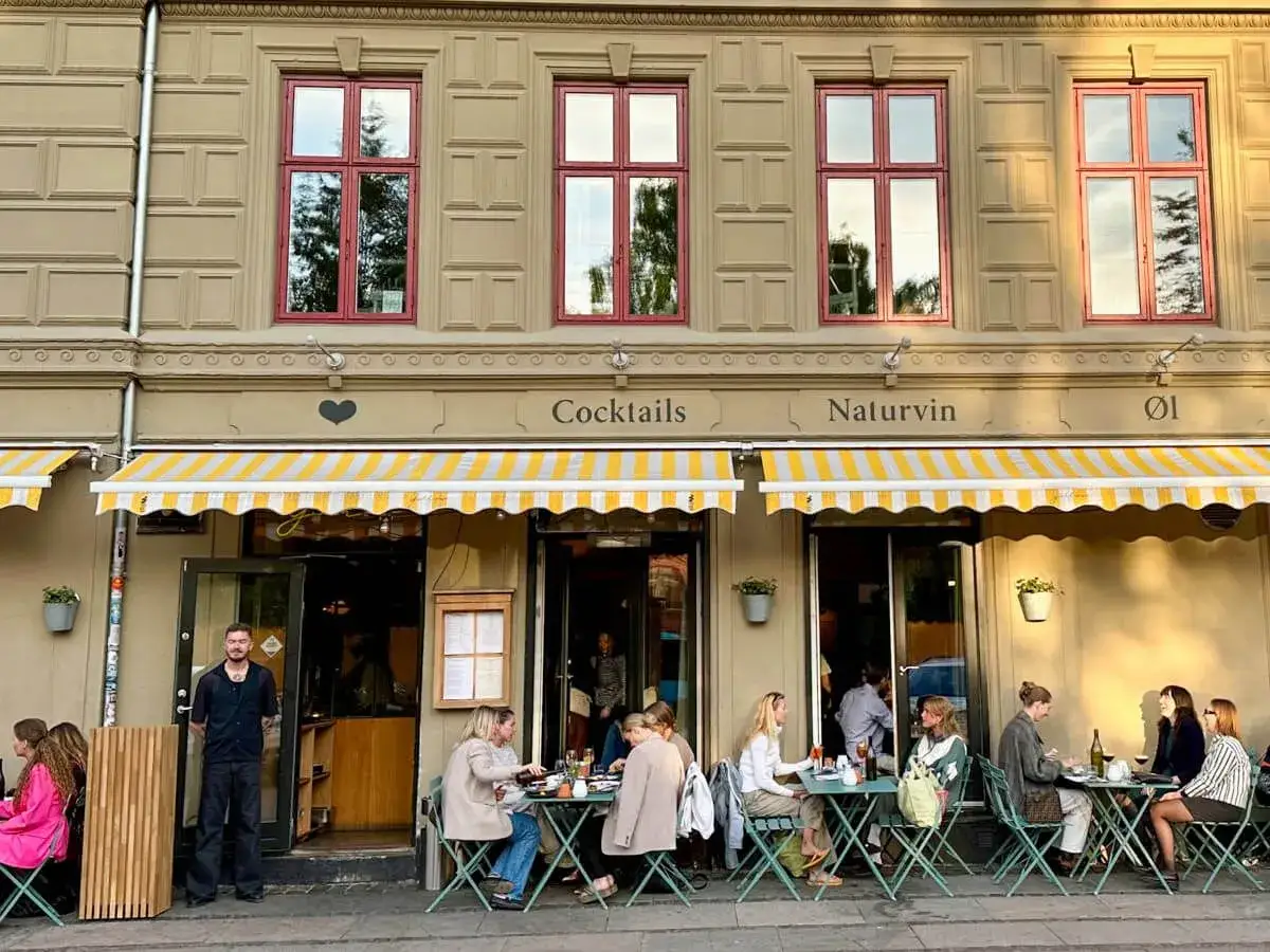 Menschen genie&szlig;en das Kopenhagen Essen auf einer Terrasse vor einem Restaurant mit gelb-wei&szlig;en Markisen.