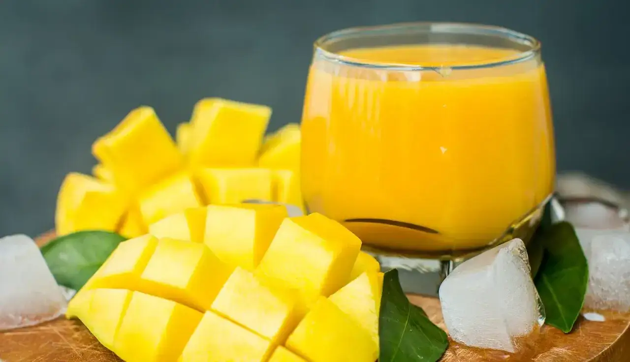 Orzeźwiający mango lassi w szklance, obok pokrojonego w kostkę soczystego mango, kostek lodu i zielonych listków.