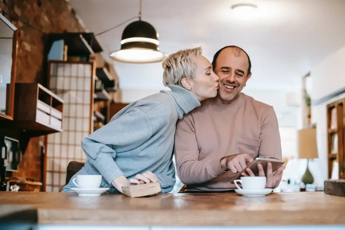 Un couple partage un moment tendre. Elle l'embrasse sur la joue, un livre et une tasse de caf&eacute; &agrave; proximit&eacute;. Ils semblent apprendre &agrave; se conna&icirc;tre, profitant de leur temps ensemble.