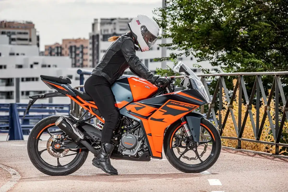 Mujer con casco blanco y chaqueta de cuero negro sobre una KTM RC 390 naranja y gris, lista para la aventura.