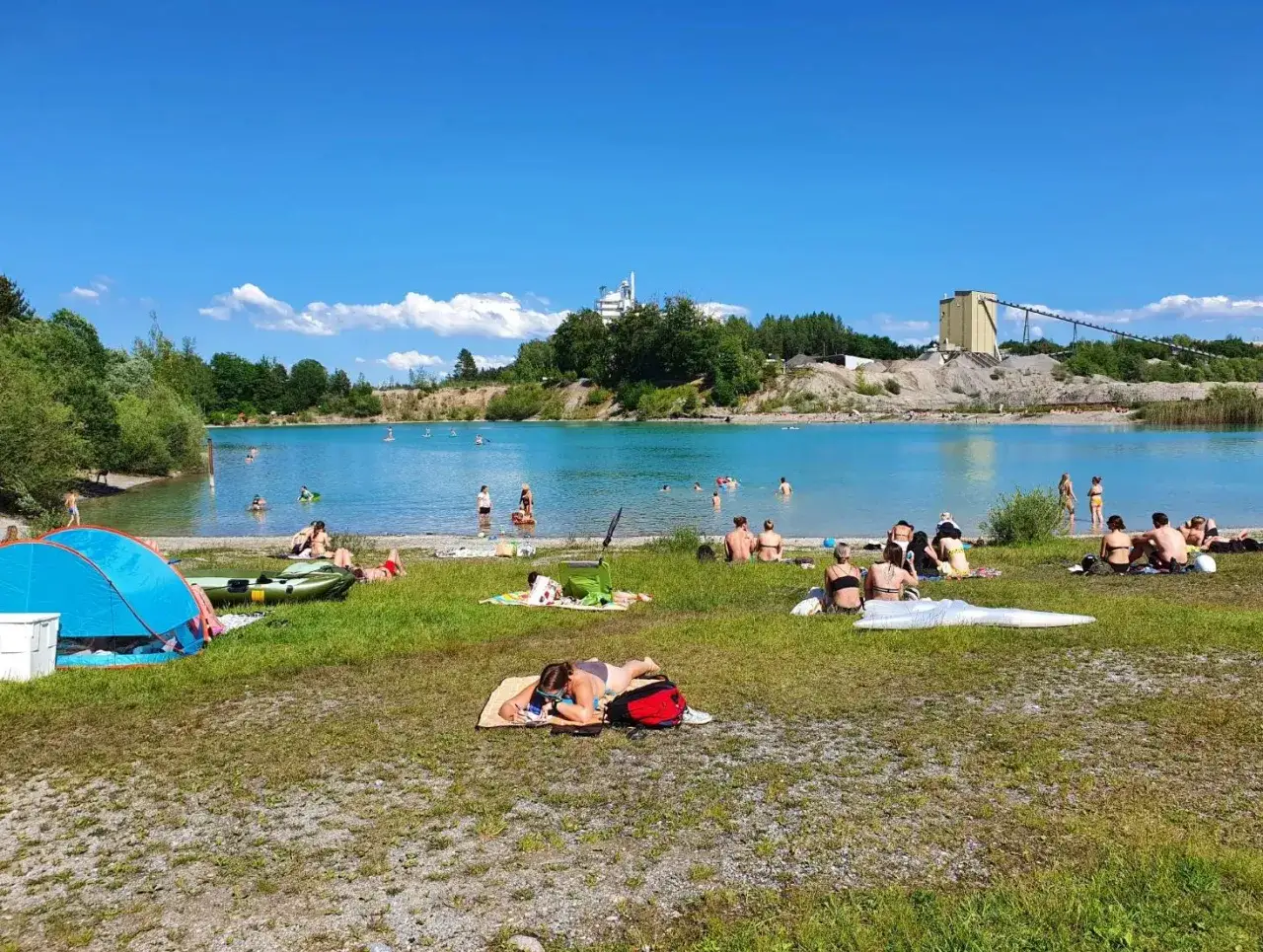 Sonniger Tag am Baggersee Grenis. Menschen entspannen am Ufer, baden im t&uuml;rkisfarbenen Wasser und genie&szlig;en die Aussicht auf die Industrieanlage.
