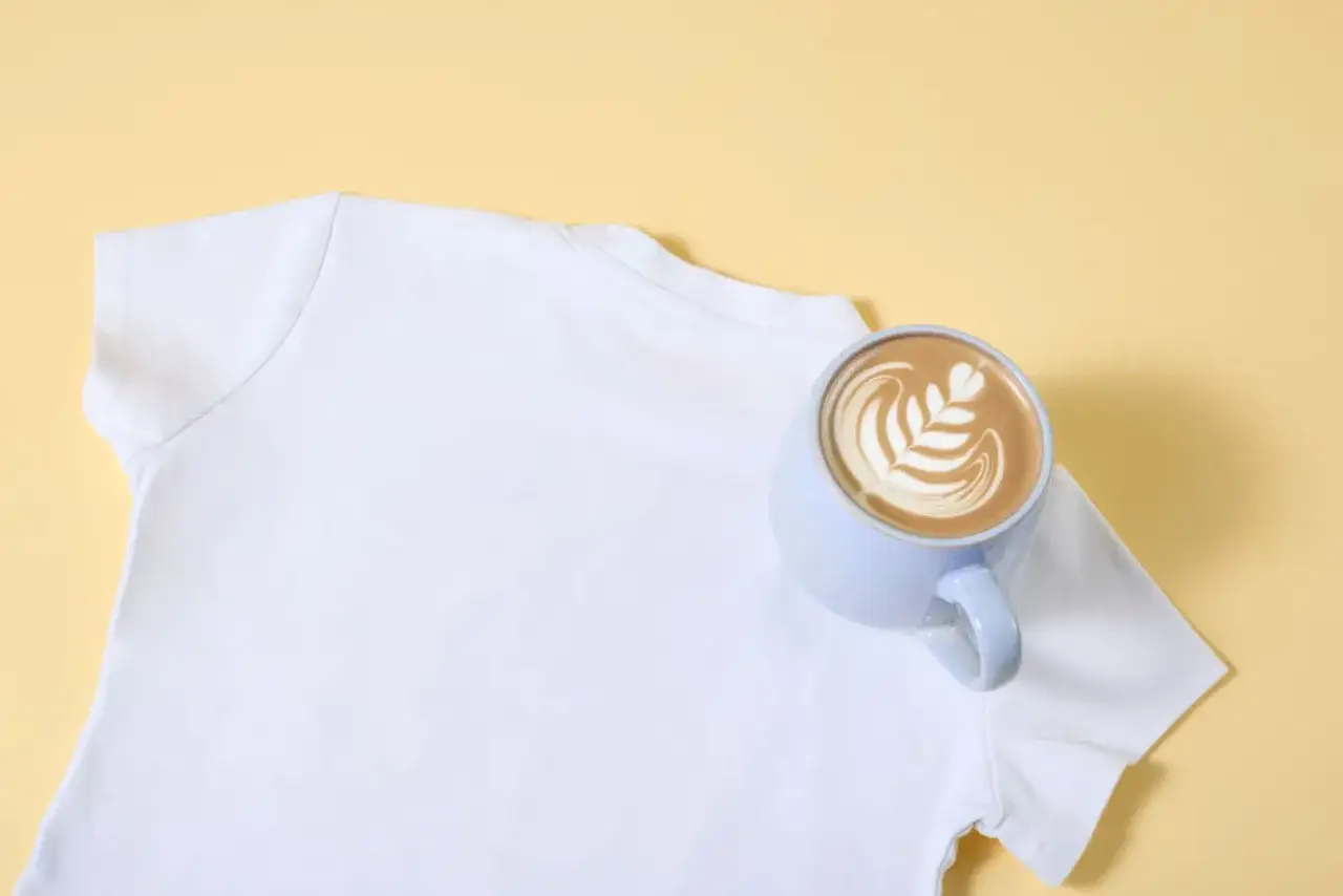Ein wei&szlig;es T-Shirt neben einer Tasse Latte Art. Ein kleiner Unfall mit Kaffee kann passieren, aber kaffeeflecken entfernen ist m&ouml;glich.