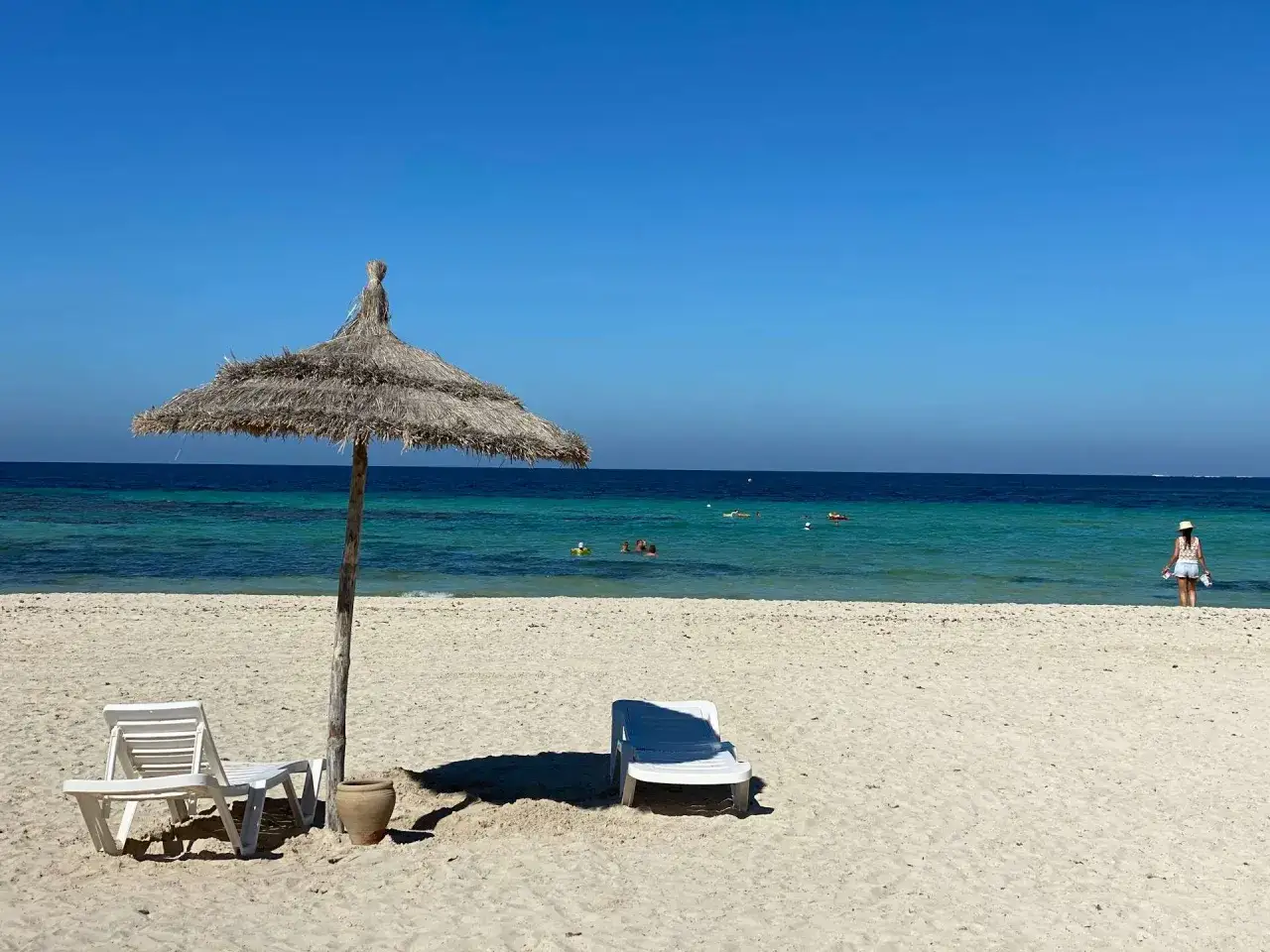 Słoneczna plaża na Djerbie, z leżakami i parasolem. W wodzie kąpią się ludzie, a na brzegu stoi kobieta. Czy Djerba jest bezpieczna dla kobiet? Tak, to popularny kierunek turystyczny.