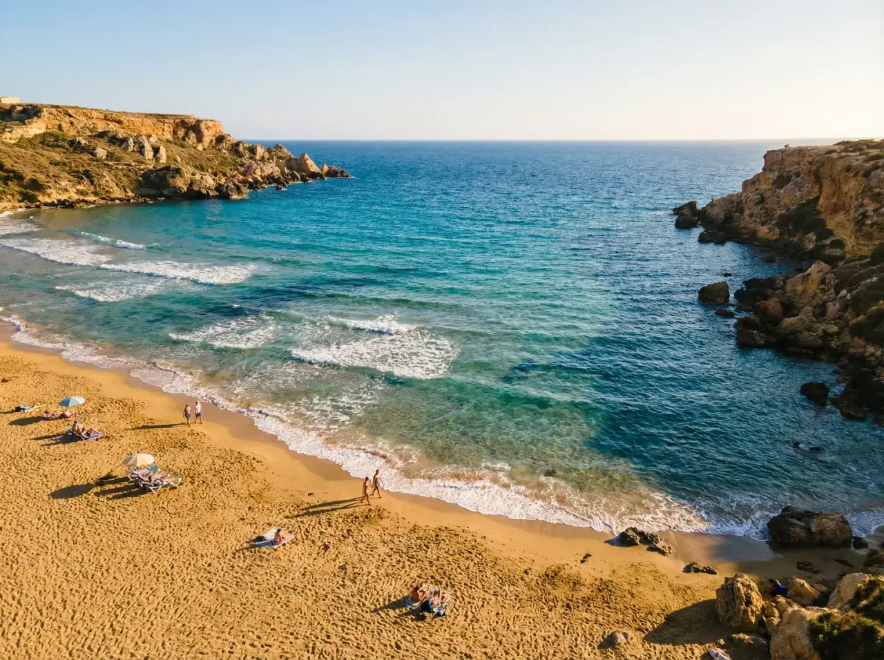 En gyllene strand vid Golden Bay, Malta, med turkost vatten och klippor. Några badgäster njuter av solen.