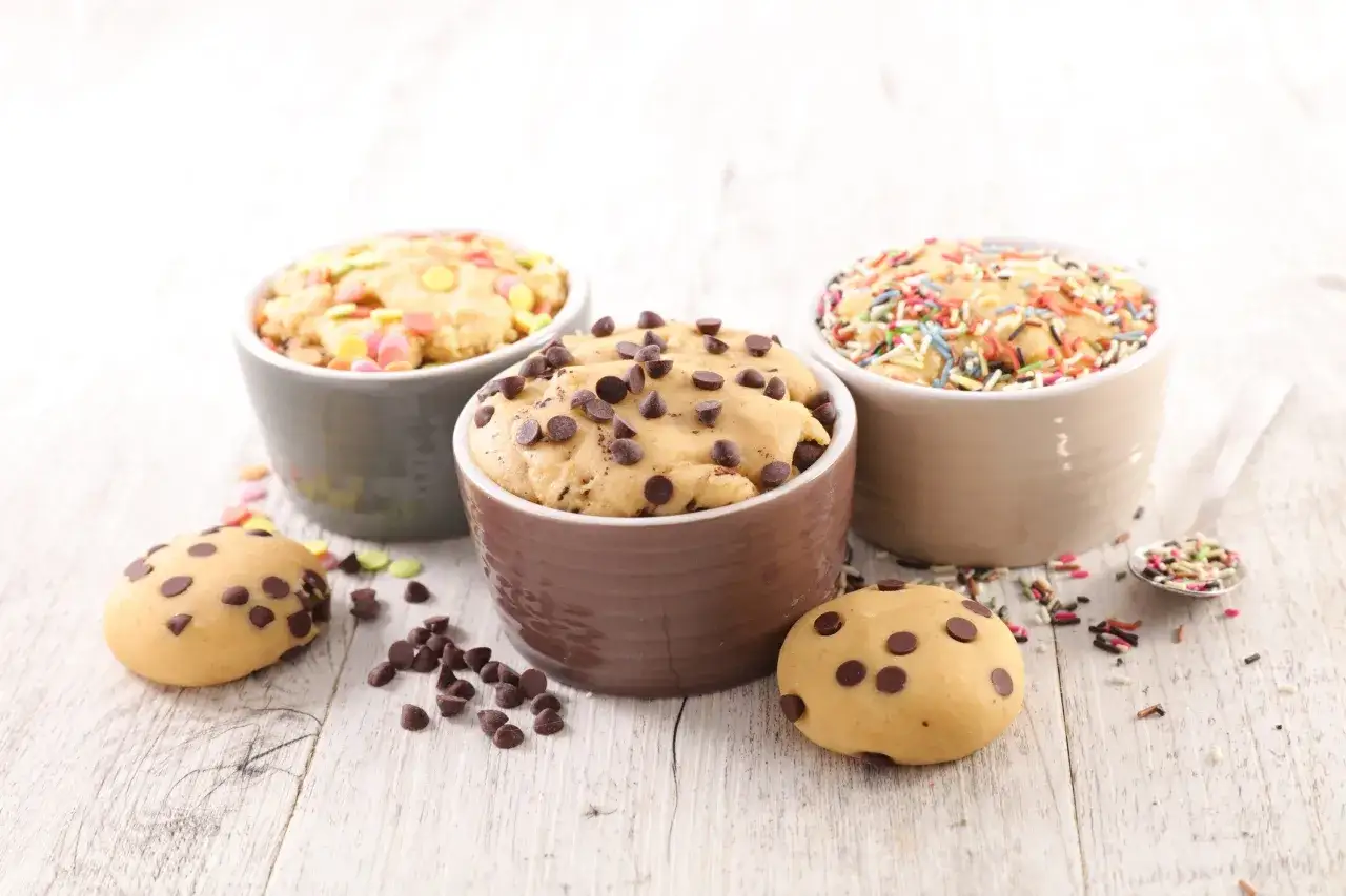 Drei Schalen mit cookie teig ohne ei, verziert mit Schokost&uuml;ckchen und bunten Streuseln, daneben zwei Kekse.