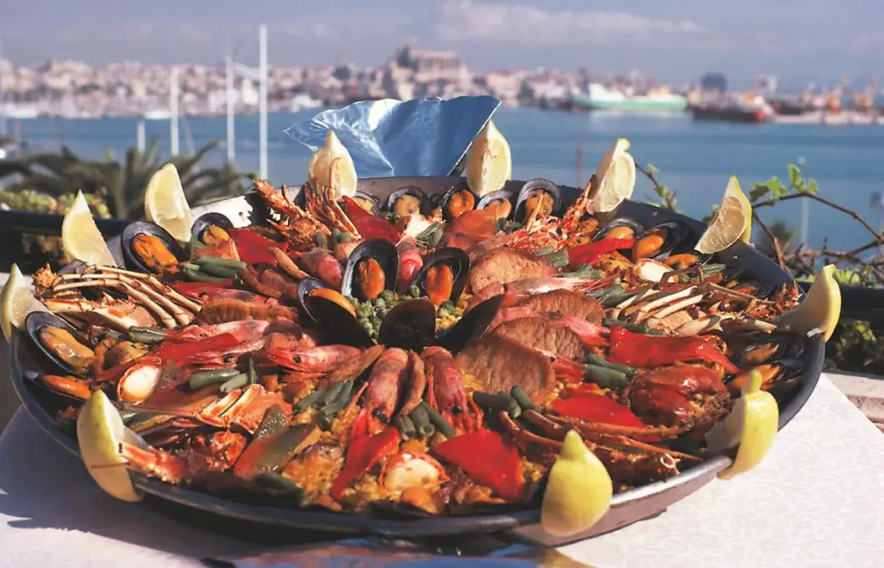 Pyszna paella, kwintesencja kuchni hiszpańskiej, z krewetkami, małżami i homarami, ozdobiona cytryną, na tle portu.