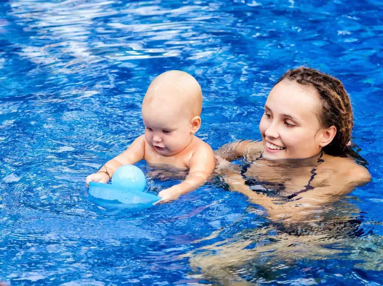 Uśmiechnięta mama i niemowlę bawią się w basenie z niebieską zabawką.