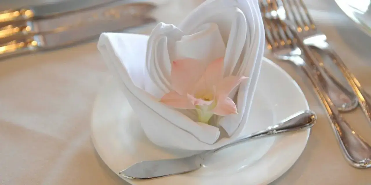 Un élégant noeud serviette mariage blanc orné d'une fleur rose pâle, sur une assiette avec des couverts argentés.