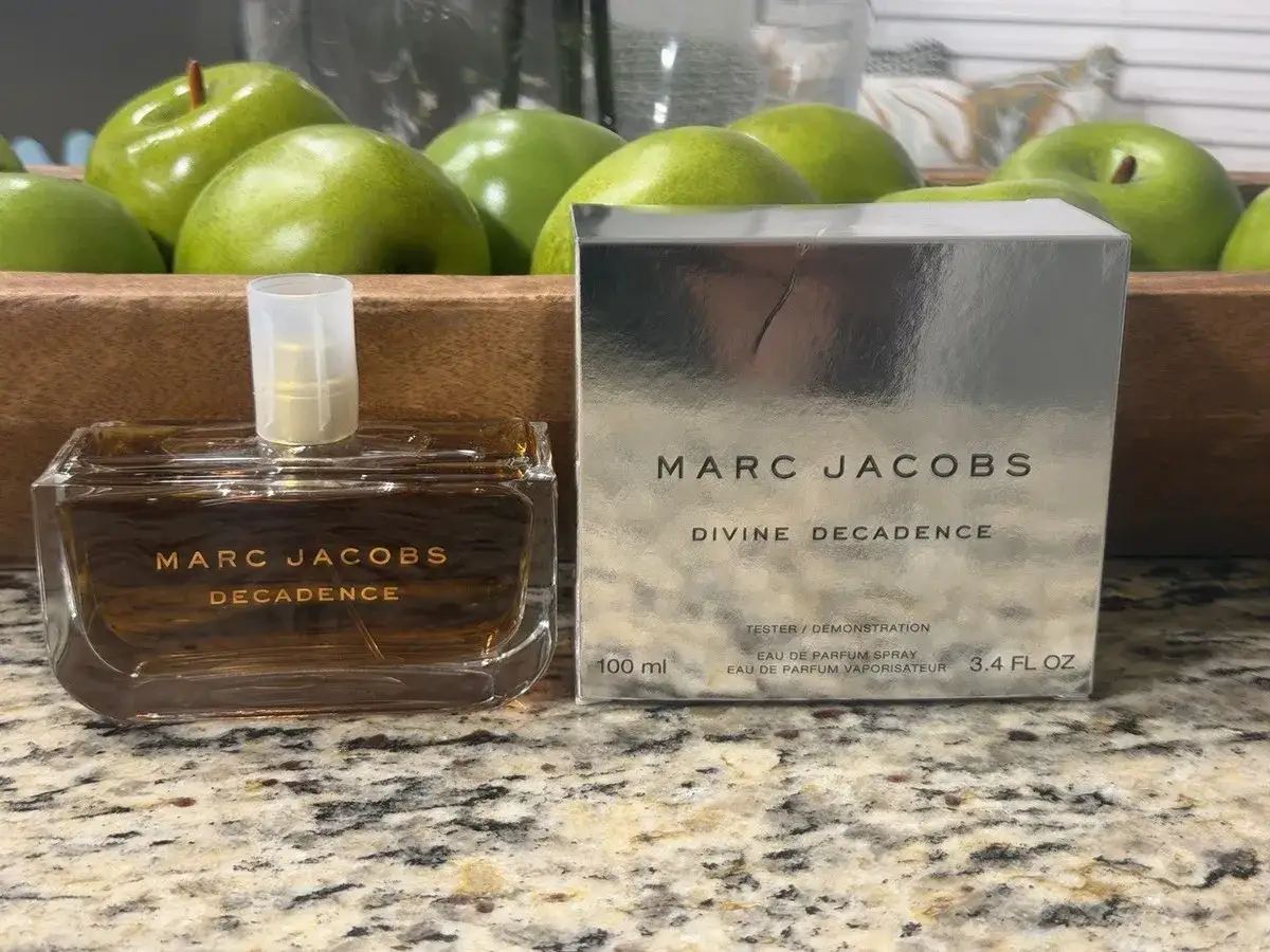 Flakon perfum Marc Jacobs Decadence i jego srebrne pudełko na tle zielonych jabłek.