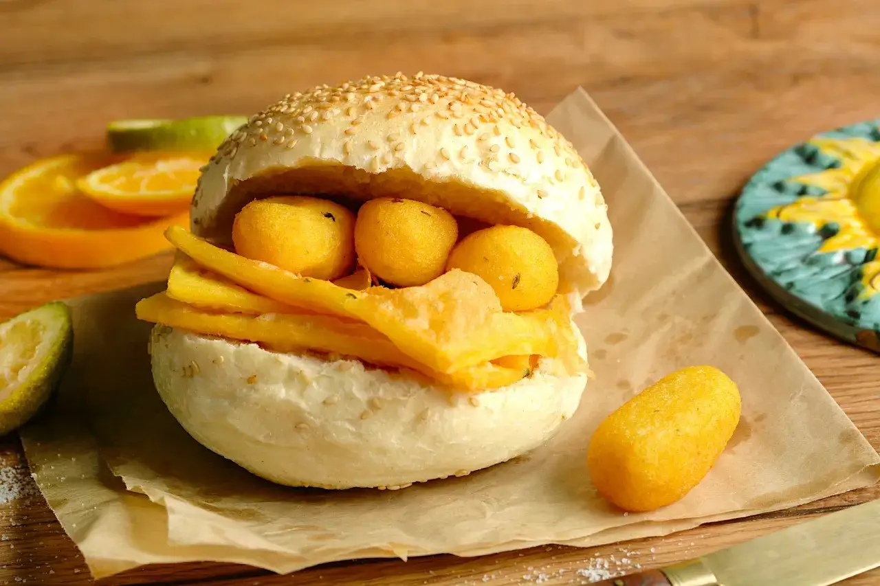 Panino con pane e panelle fritte, accompagnato da spicchi d'arancia e limone su carta da forno.