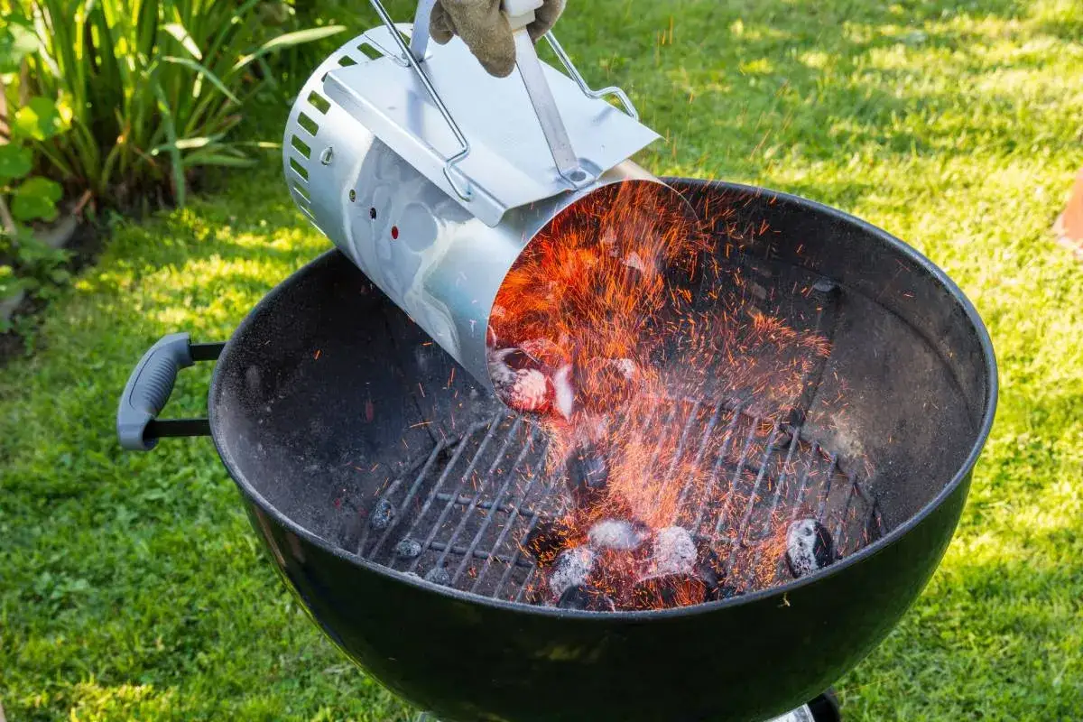 Manos enguantadas vierten brasas incandescentes de un encendedor de chimenea a una barbacoa de carb&oacute;n, mostrando c&oacute;mo encender una barbacoa de carb&oacute;n.