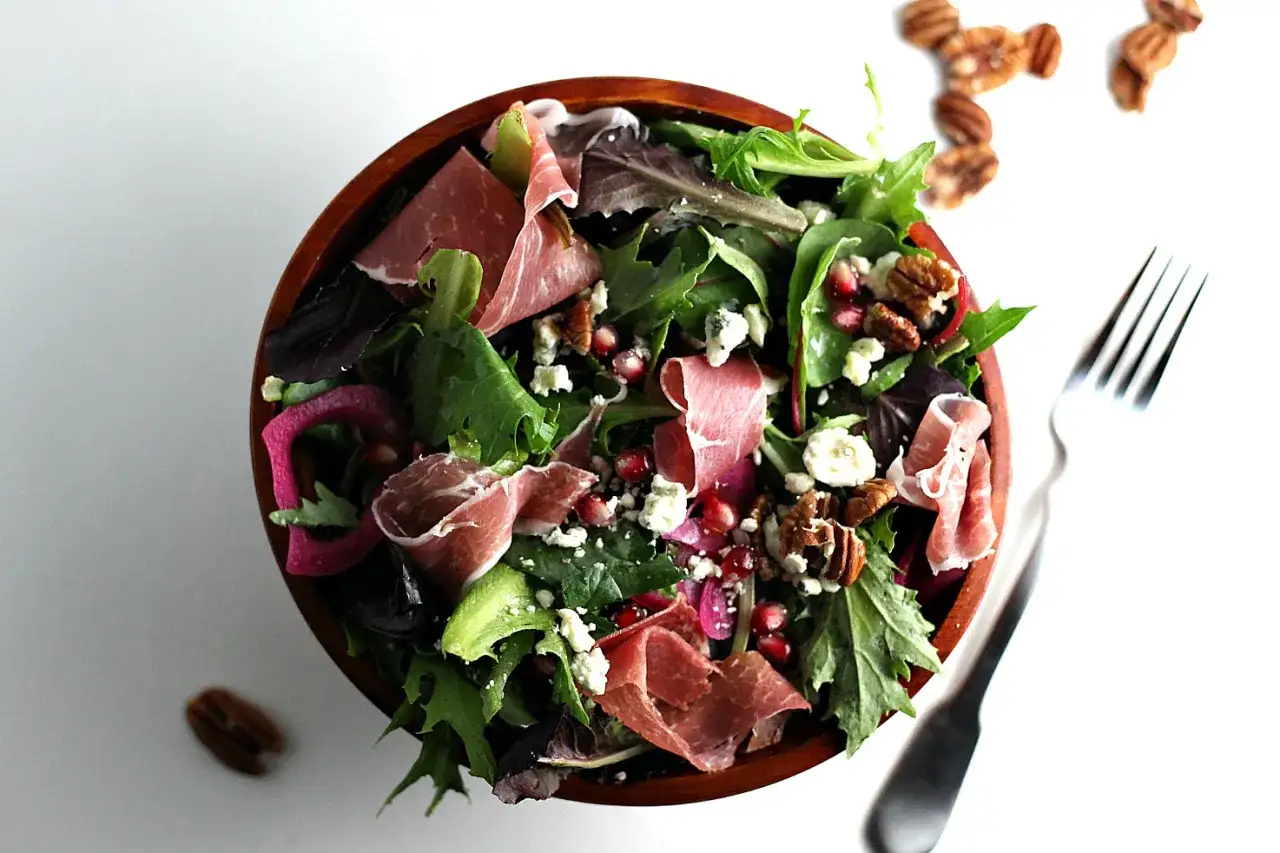 En härlig sallad med prosciutto, granatäpple, nötter och blåmögelost i en träskål. En gaffel ligger bredvid.