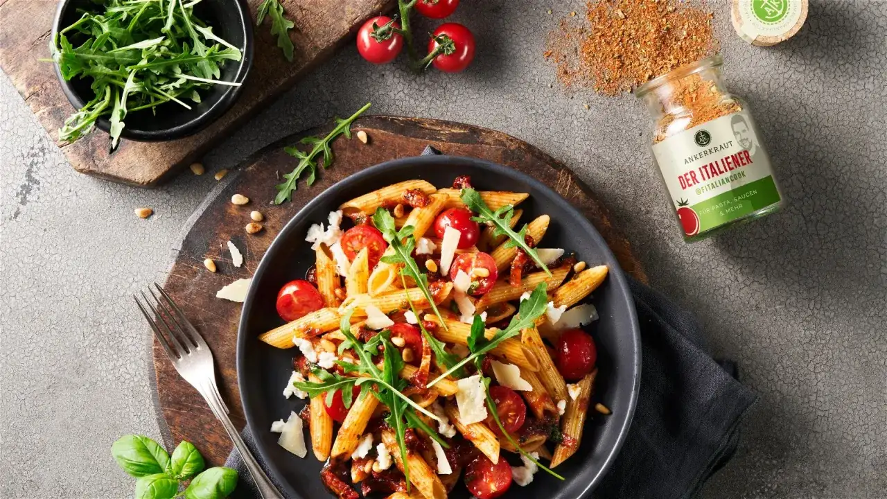 Ein köstlicher nudelsalat mit getrockneten tomaten, garniert mit Rucola und Pinienkernen. Daneben ein Glas Gewürz