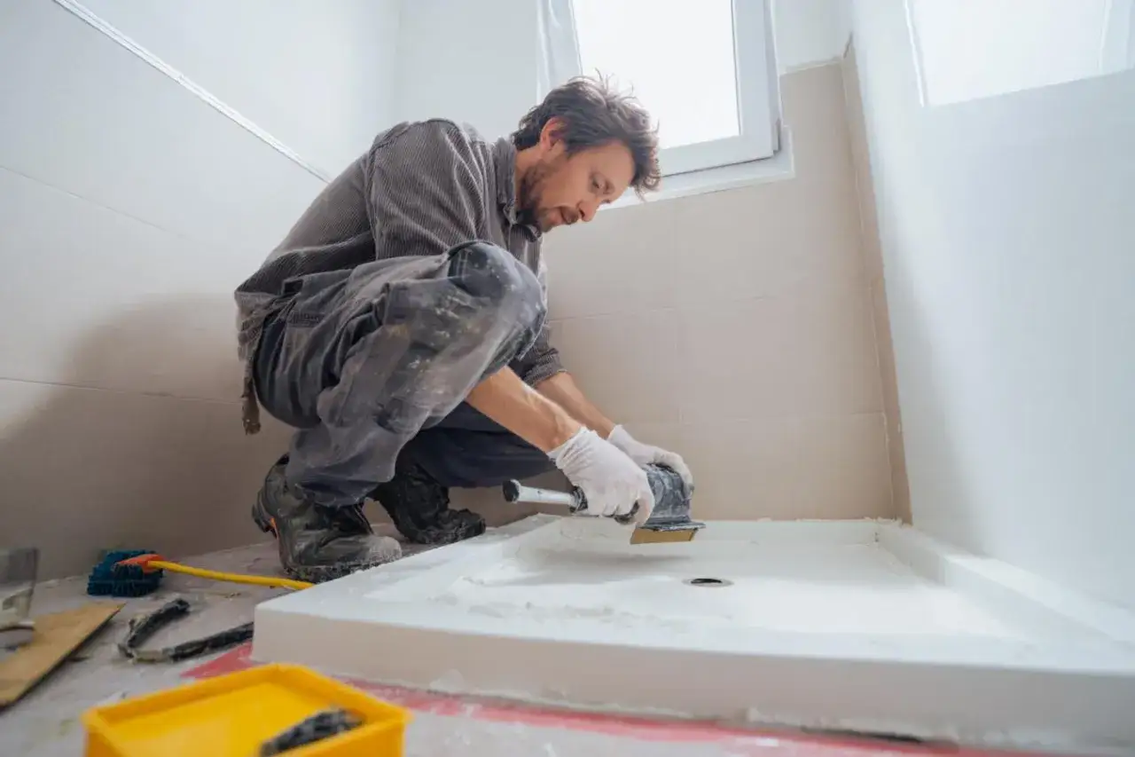 Un homme pr&eacute;pare le sol pour installer un receveur de douche. Il utilise une ponceuse pour lisser la surface, une &eacute;tape cl&eacute; pour savoir avec quoi coller un receveur de douche.