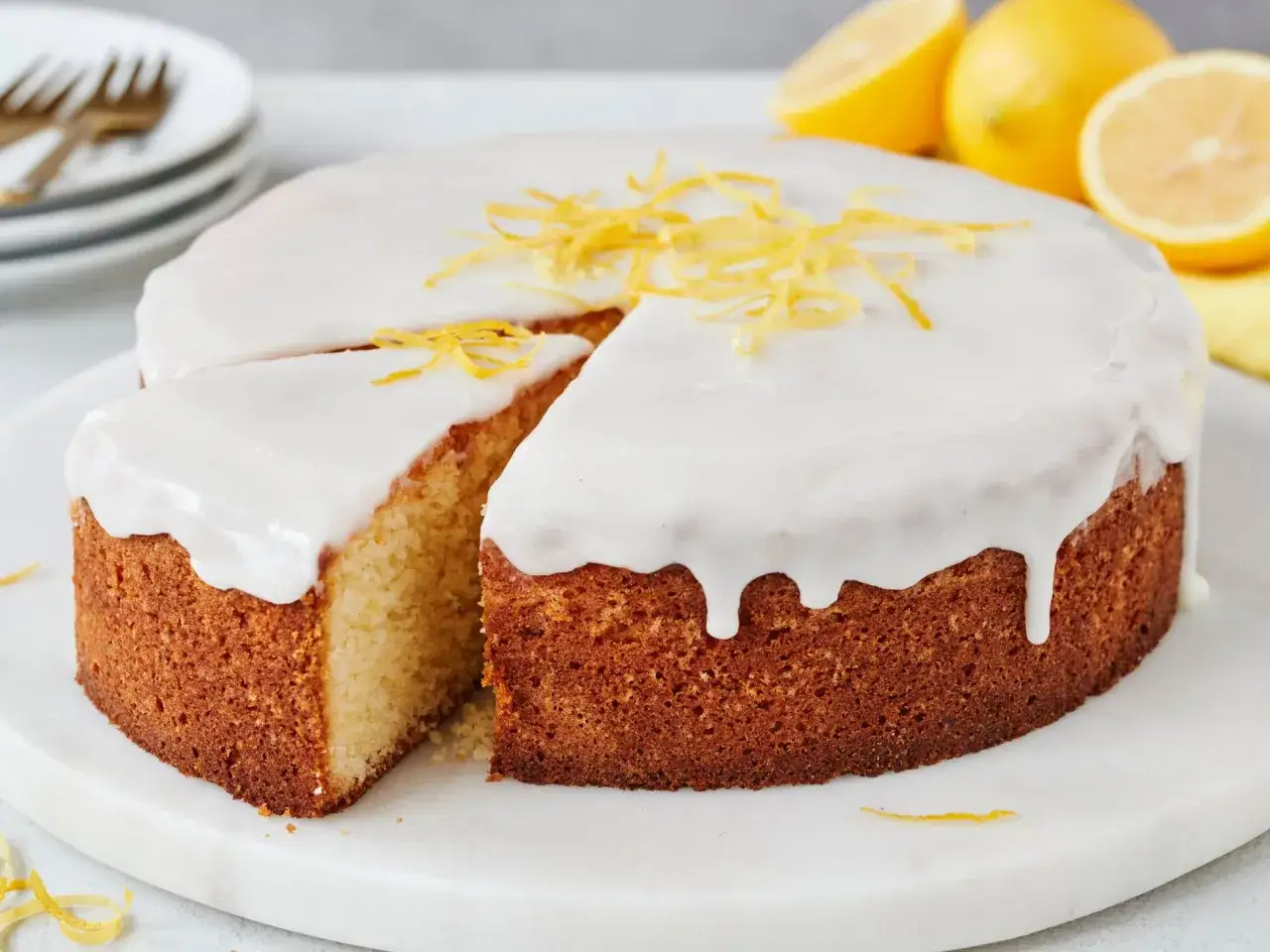 Ein saftiger sizilianischer Zitronenkuchen mit wei&szlig;er Glasur und Zitronenzesten. Daneben liegen frische Zitronen.