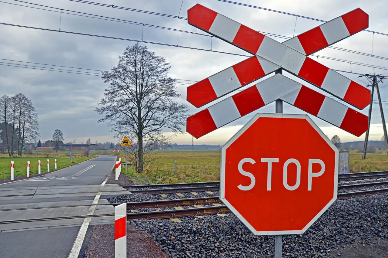 Przed przejazdem kolejowym stoją zapory kolejowe i znak STOP. Droga prowadzi przez tory.
