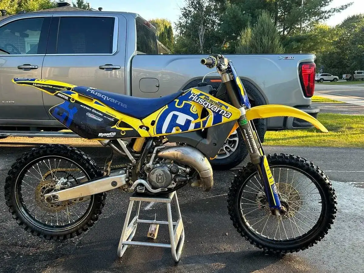Żółto-niebieski motocykl Husqvarna CR 125 na stojaku, obok pickup'a. Dane techniczne tego modelu są imponujące.