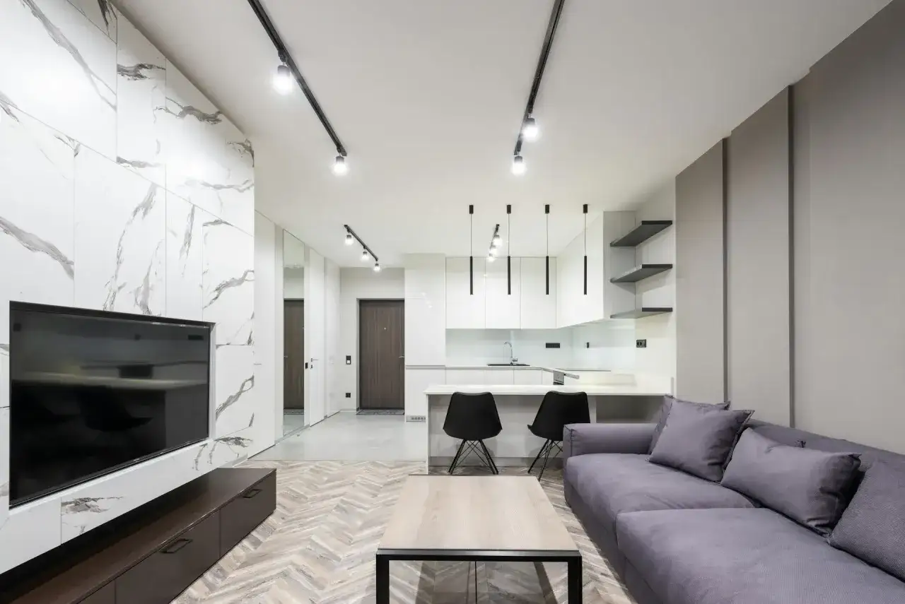 Nowoczesny salon z aneksem kuchennym. Montaż szynoprzewodów z reflektorami podkreśla minimalistyczny design.
