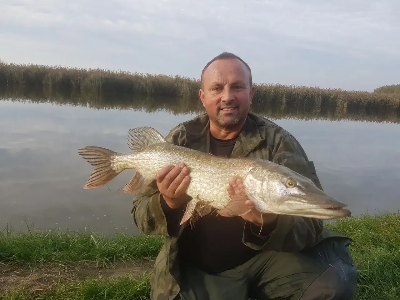 Szczęśliwy wędkarz z okazałym szczupakiem na tle spokojnego łowiska Stary San Hurko.