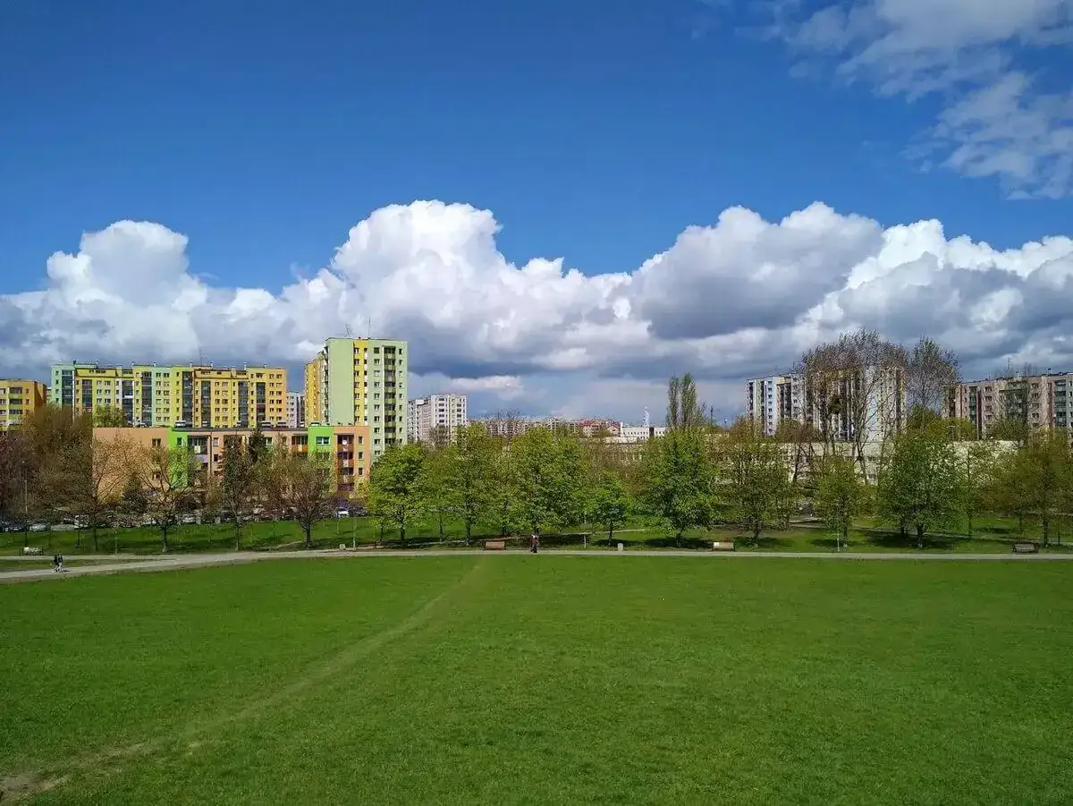 Zielona przestrzeń w górczewska park, z kolorowymi blokami mieszkalnymi w tle i błękitnym niebem z białymi chmurami.