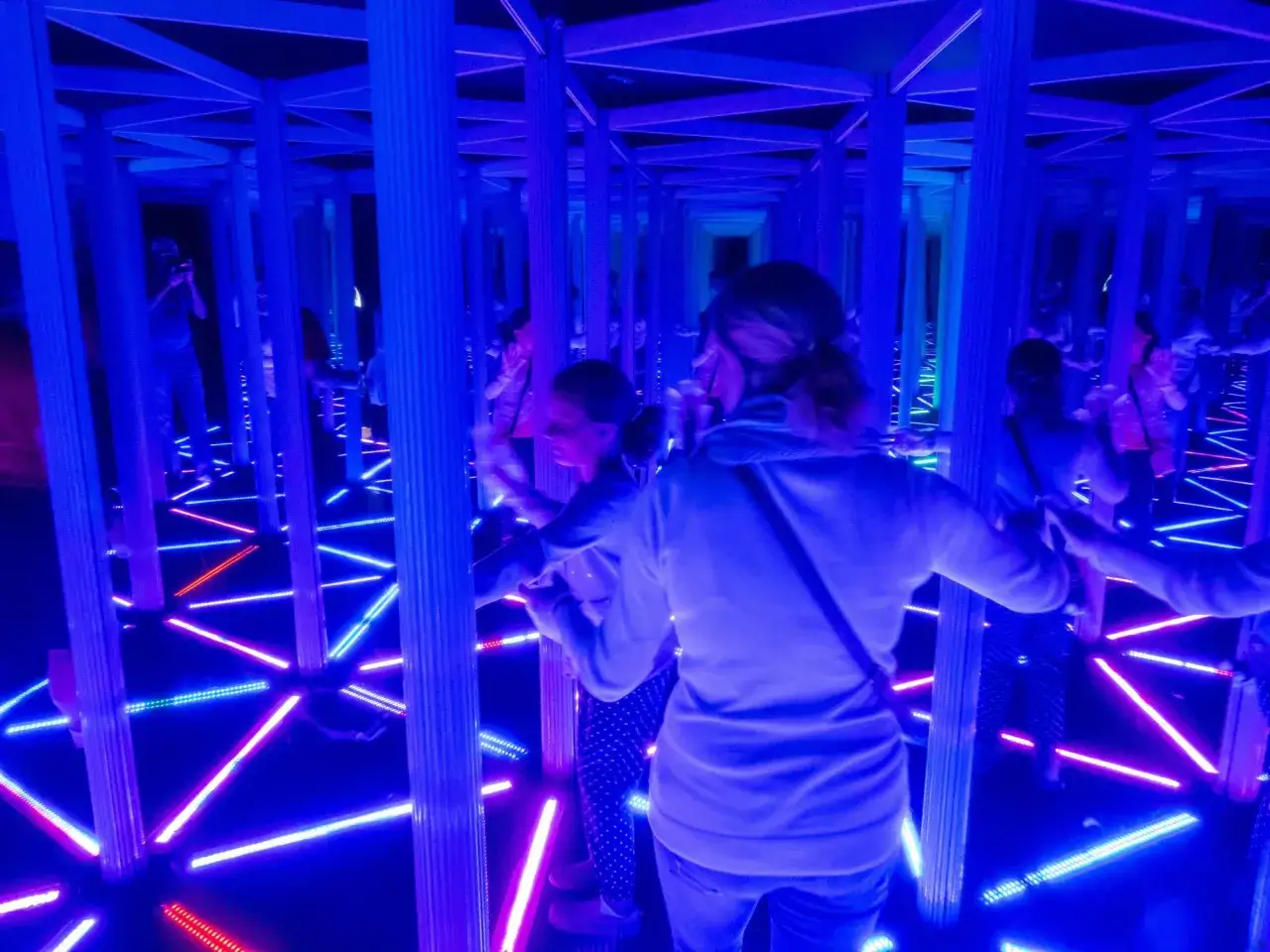 Wewnątrz house of attractions, ludzie poruszają się wśr&oacute;d luster i neonowych świateł, tworząc iluzję nieskończoności.