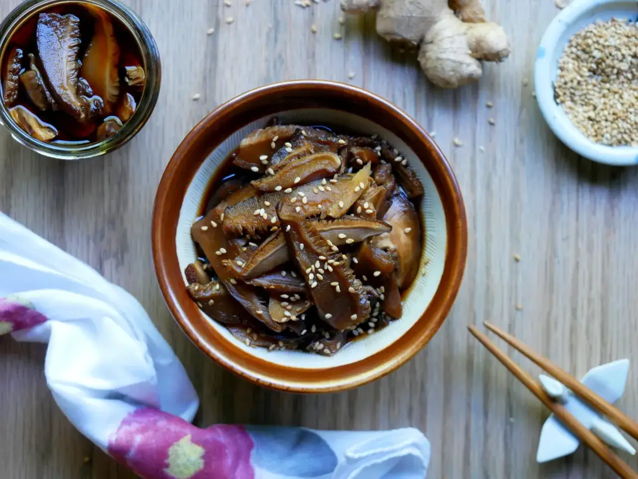 A bowl of glistening pickled shiitake mushrooms, sprinkled with sesame seeds, sits on a wooden surface. A jar of more mushrooms, ginger, sesame seeds, and chopsticks are nearby.