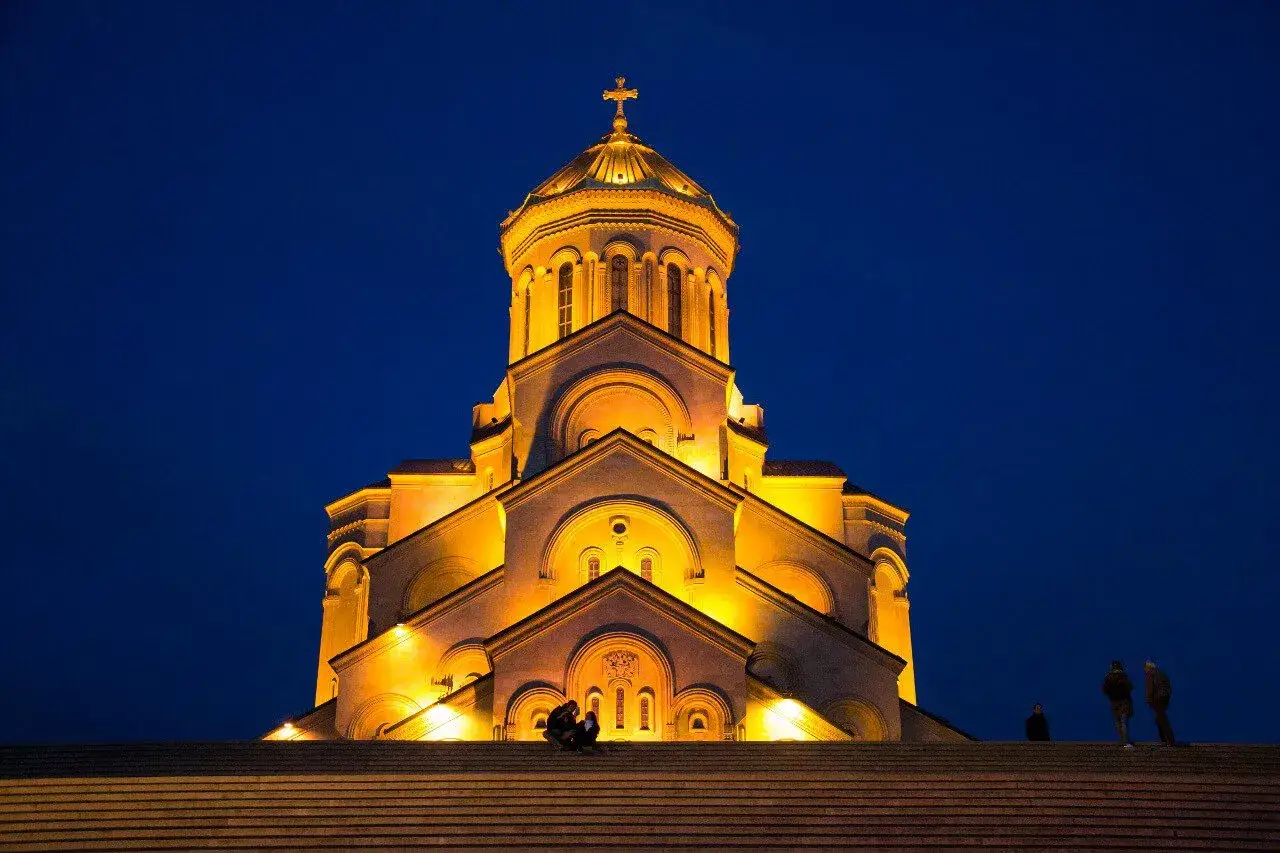 Katedra w Tbilisi, stolicy państwa Gruzja, oświetlona nocą. Kilka osób siedzi na schodach.