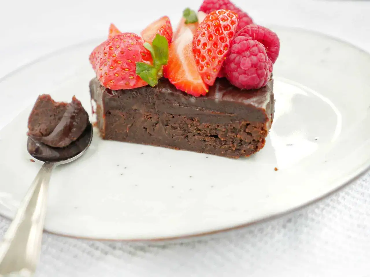 En bit chokladbrownie toppad med f&auml;rska jordgubbar och hallon. En sked med brownie-smet bredvid. Perfekt f&ouml;r ett brownie recept.