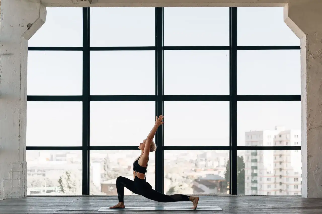 Kvinna i svart träningsklädsel utför en jivamukti yoga-position framför ett stort fönster med utsikt över staden.