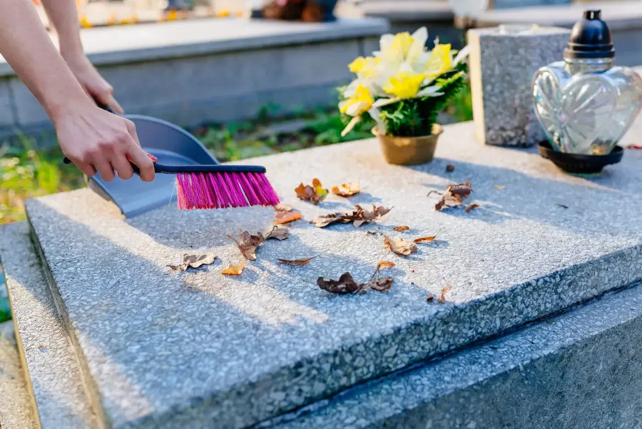 Ręce sprzątają grób przed Świętem Zmarłych, usuwając liście. Harmonogram prac obejmuje też kwiaty i znicze.