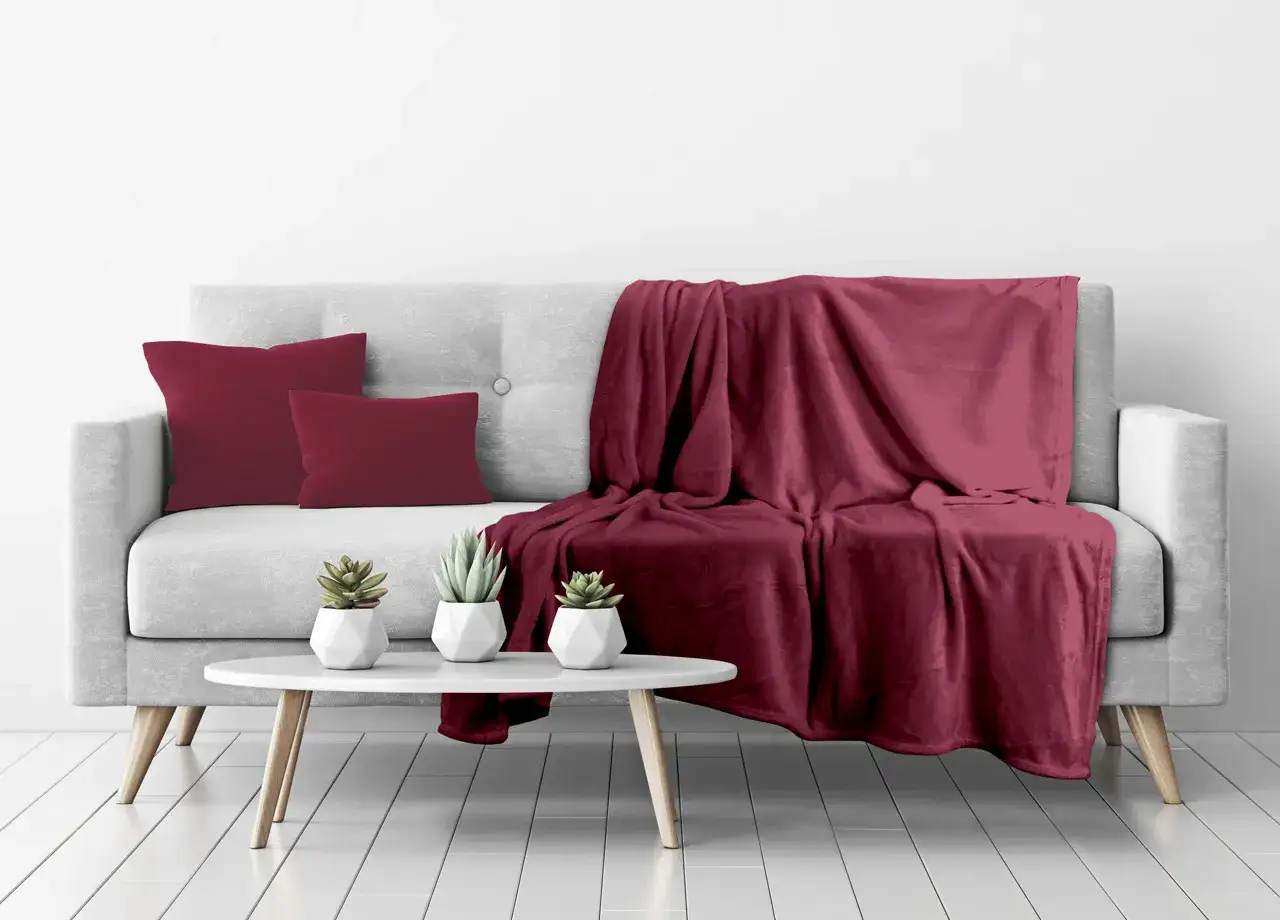 Un morbido plaid bordeaux copre un divano grigio, con cuscini coordinati e piccole piante sul tavolino.